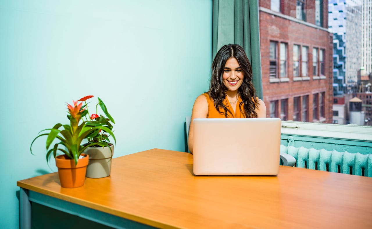 gallery-1 Confident woman in a bright office working on a laptop, surrounded by urban scenery and greenery.
